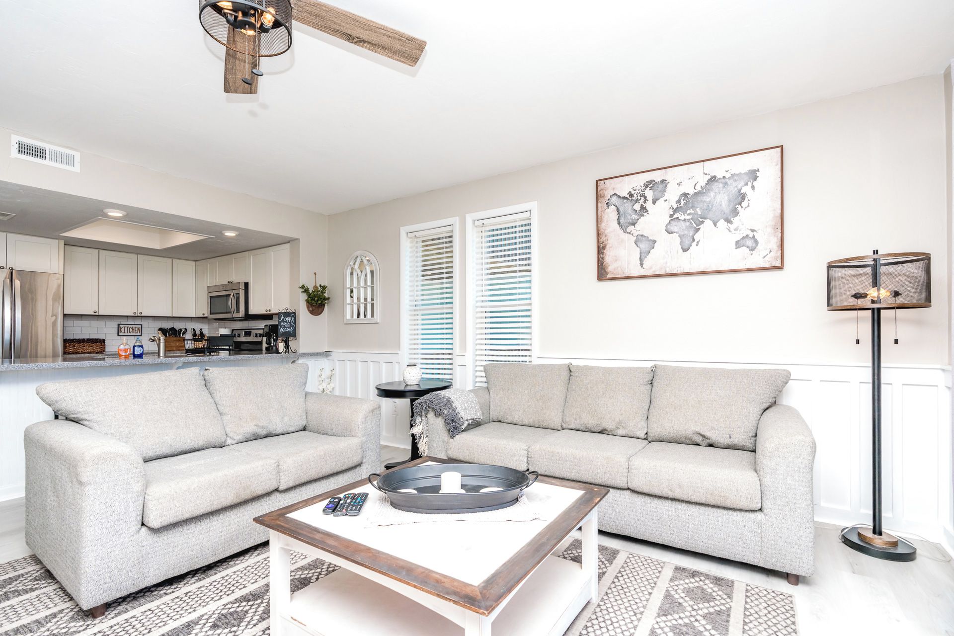 A living room with a grey sofa, a coffee table, a world map on the wall, and a kitchen in the background.