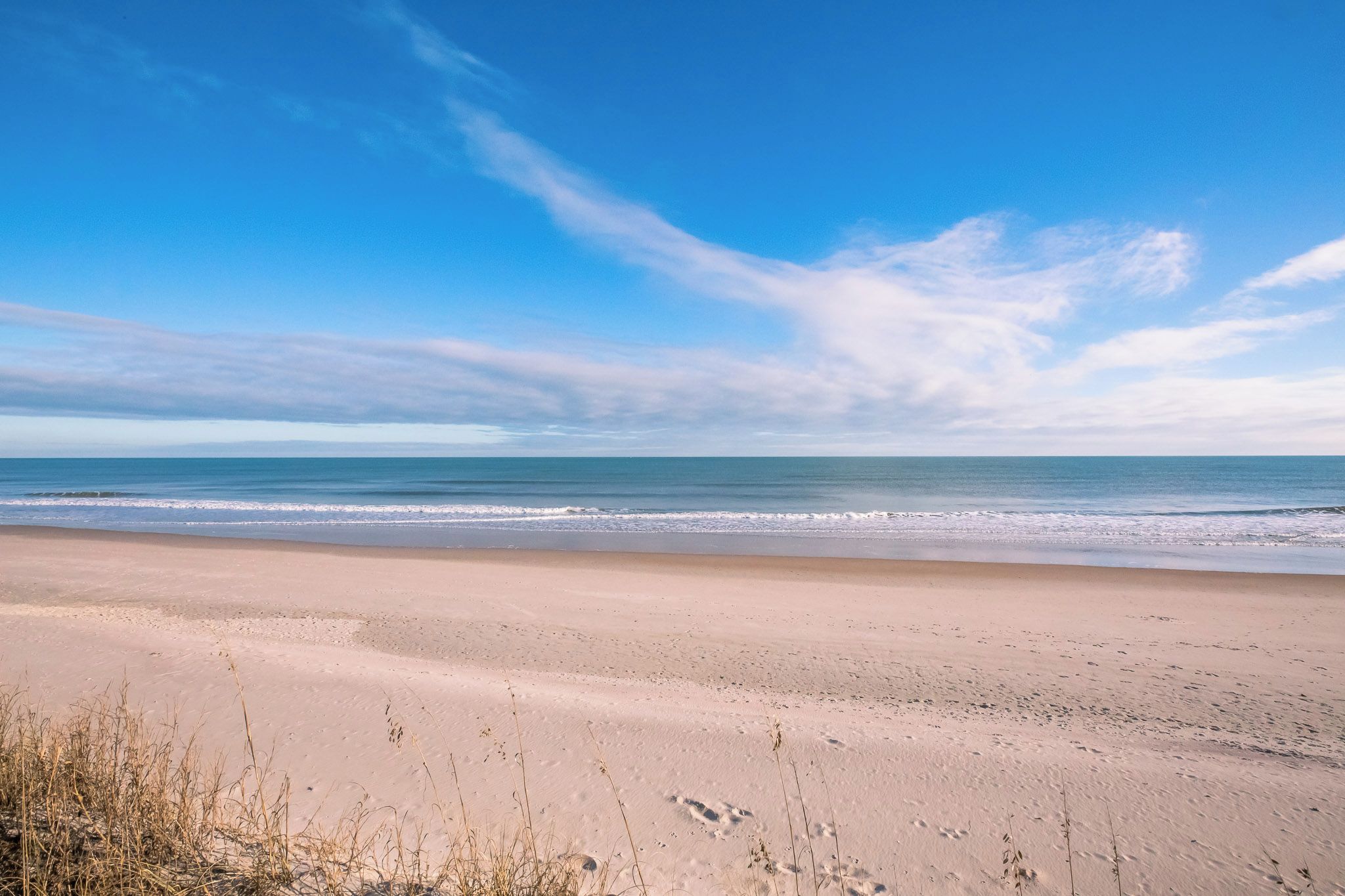A wide sandy beach with gentle waves, clear sky, and sparse vegetation.