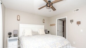Quarto com cama, mesas de cabeceira, ventilador de teto e porta para outro espaço.