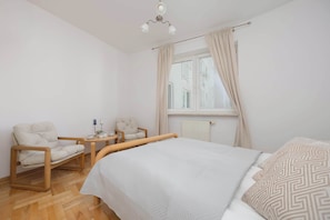A bedroom with a bed, two chairs, a small table, a window with curtains, and a radiator. - Spacious Apartment with Balcony | Parking and 400 m to Metro (Warszawa)