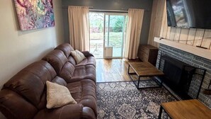 A living room with a brown leather sofa, a flat-screen TV mounted above a fireplace, and a large abstract painting on the wall. - Backyard Escape + Hot Tub - Minutes to Red Rocks & Denver-Large Basement Hangout (Arvada)