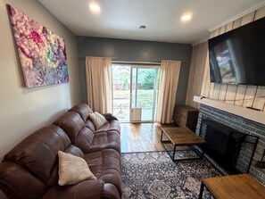 A living room with a brown leather sofa, a flat-screen TV mounted above a fireplace, and a large abstract painting on the wall. - Backyard Escape + Hot Tub - Minutes to Red Rocks & Denver-Large Basement Hangout (Arvada)