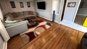 A living room with a sofa, a flat-screen TV, wooden flooring, and a window with blinds. - Backyard Escape + Hot Tub - Minutes to Red Rocks & Denver-Large Basement Hangout (Arvada)