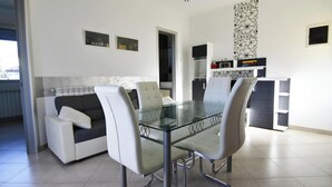 Un comedor moderno con una mesa y sillas de vidrio, un sofá con un cojín y una cocina al fondo.