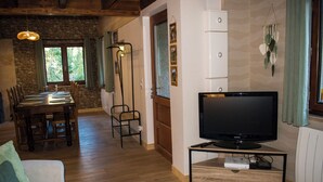 A living room with a television on a stand, a dining table with chairs, a sofa, and a stone wall. - Le P'tit Chalet, at 1000m altitude (Campan)