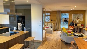 Eine moderne Küche mit einer zentralen Kochinsel, ein Essbereich mit einem Holztisch und Barhocker, eine Sitzecke mit bunten Kissen und ein großes Fenster mit Blick auf Bäume.