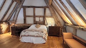 Een slaapkamer met een schuine kap, houten balken, een bed, een stoel, een nachtkastje en een bankje.