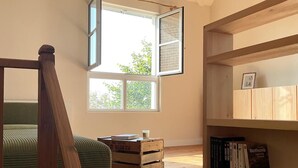 Une pièce avec un lit en mezzanine, une bibliothèque en bois, une fenêtre avec des volets ouverts et une petite table sur laquelle sont posés des livres.