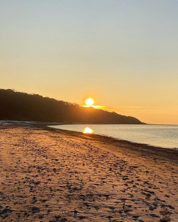 A beach with the sun setting over the water, a sandy shore, and a hilly landscape in the background.