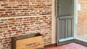 Une pièce avec un mur en briques, une malle en bois portant l’inscription « GRAMANT BRUT », un chapeau posé sur un porte-chapeau et une porte verte.