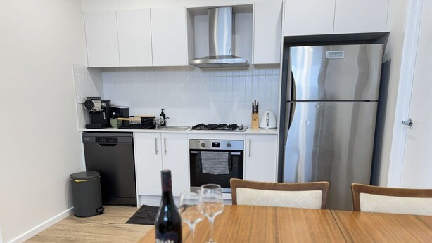Une cuisine moderne avec des meubles blancs, une hotte en acier inoxydable, un réfrigérateur et une table à manger agrémentée de verres à vin et d’une bouteille.
