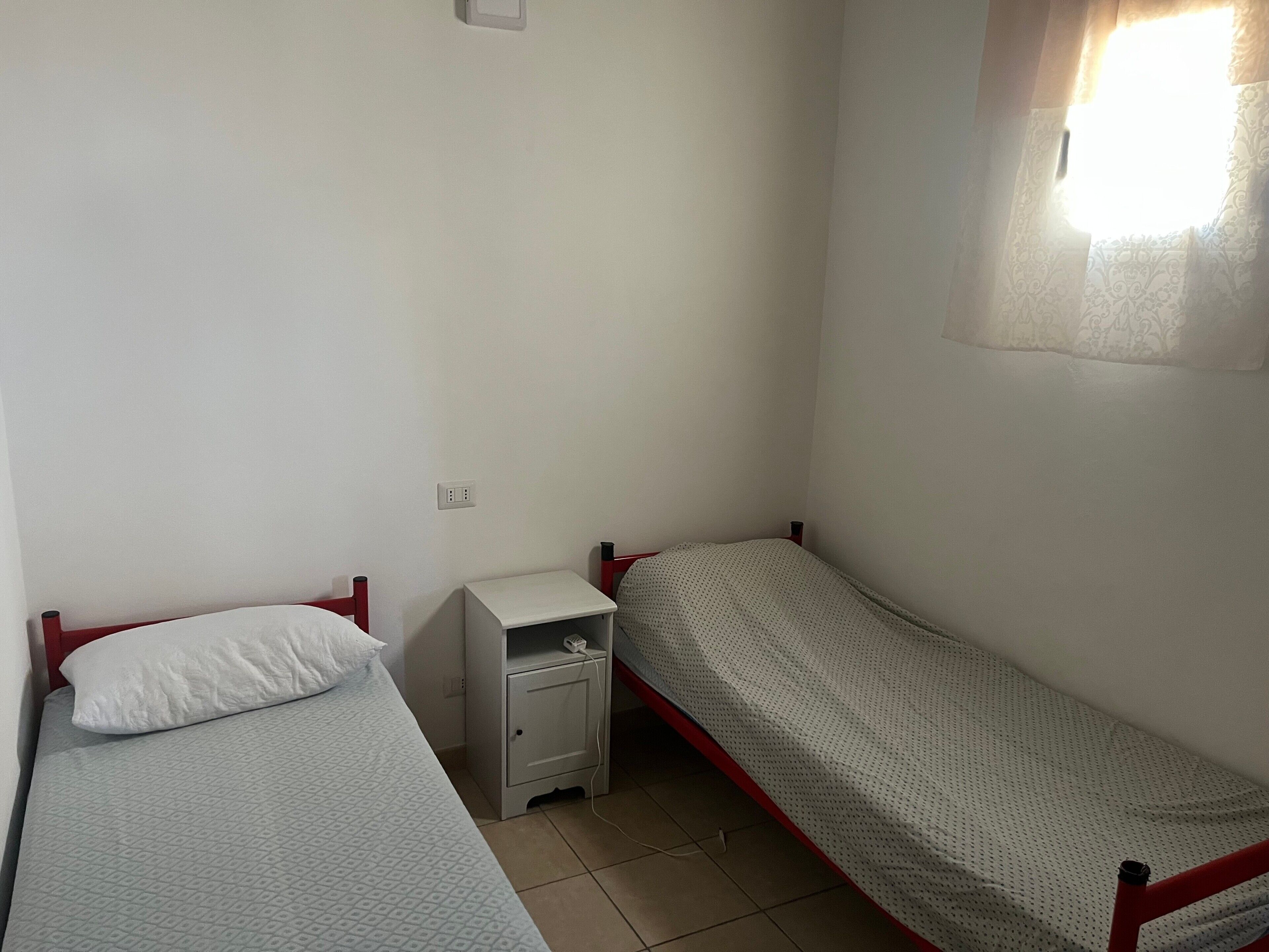 Two single beds with red frames, a small bedside table, and a power outlet on the wall.