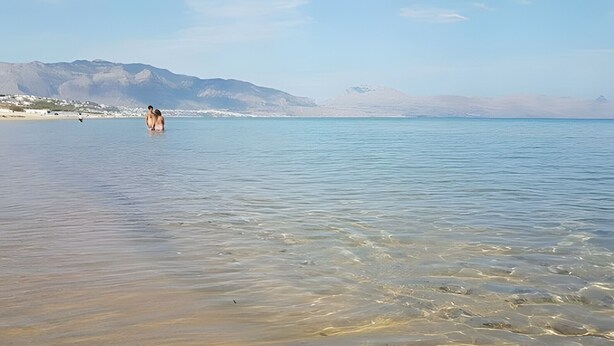 Una spiaggia con acque limpide e poco profonde, una persona in acqua e montagne sullo sfondo.