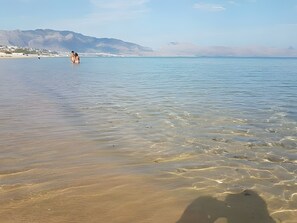 Une plage avec une eau claire et peu profonde, une personne debout dans l’eau et des montagnes en arrière-plan.