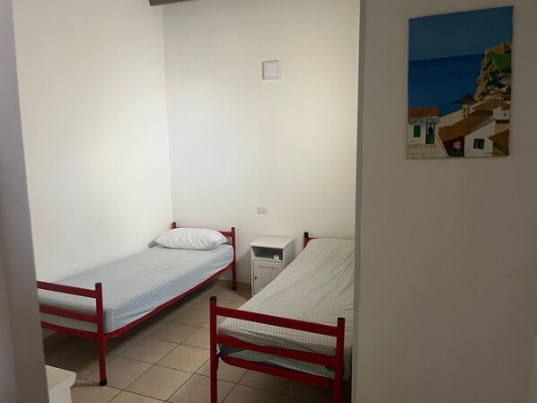 Une petite pièce, peu meublée, avec deux lits simples, une table de chevet et une prise de courant fixée au mur.