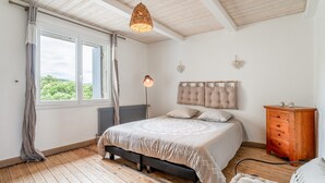 Een slaapkamer met een bed, een dressoir, een radiator, een hanglamp en een raam met gordijnen.