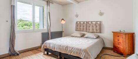 Ein Schlafzimmer mit Bett, Kommode, Heizkörper, Pendelleuchte und einem Fenster mit Vorhängen.