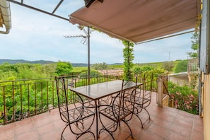Una veranda esterna coperta con tavolo e sedie, circondata da un recinto in metallo, con vista su un paesaggio verdeggiante.