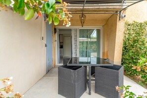 Un patio con una mesa de vidrio y sillas de mimbre, una puerta que da a una habitación, y una pared cubierta de plantas trepadoras.