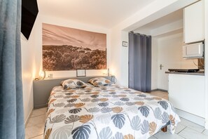 A bedroom with a bed, a large wall-mounted picture, a bedside table with a lamp, and a view of a kitchen area. - Gite Lavande – La Bastide St Jacques (Vinsobres)