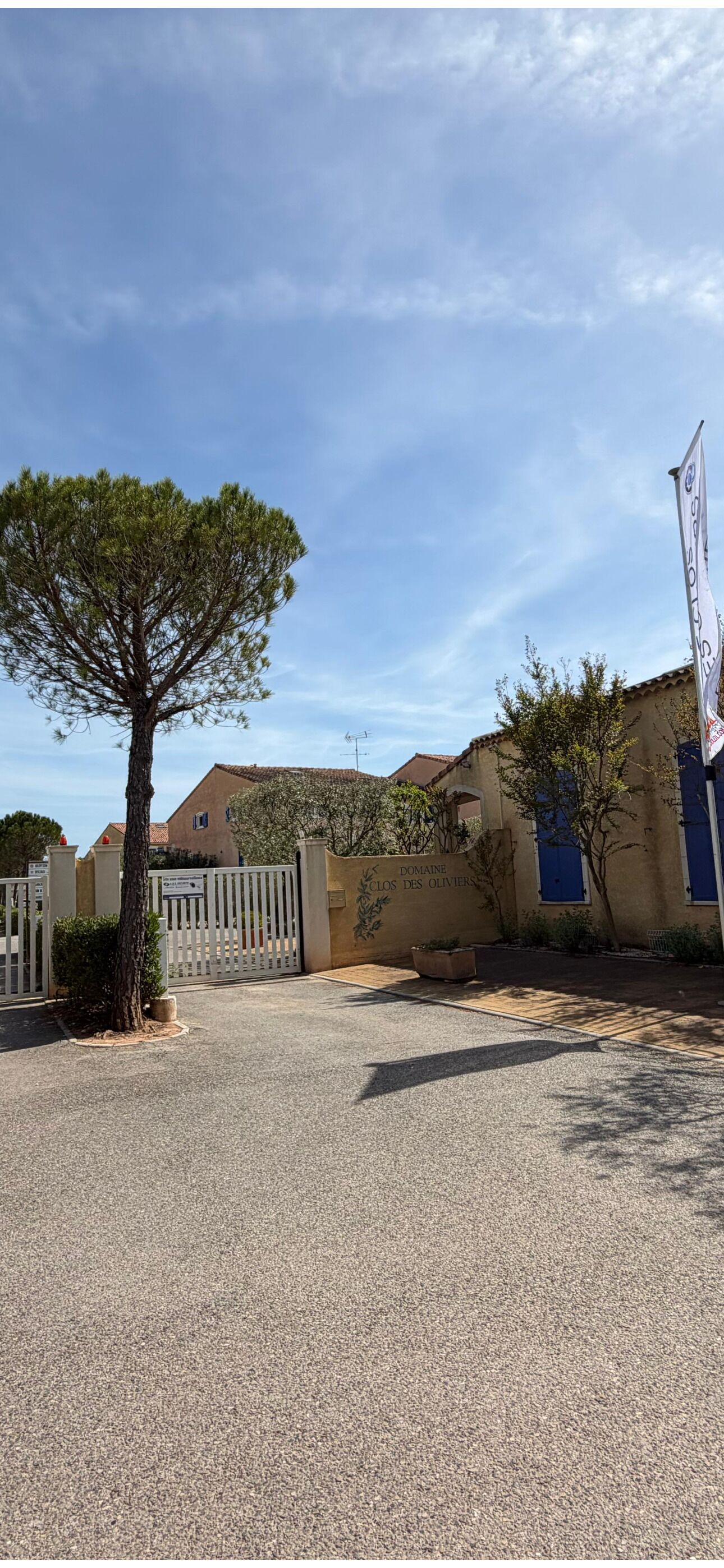 Une entrée fermée par un portail, avec un arbre, un bâtiment aux volets bleus et un drapeau.