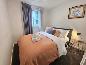 Ein ordentlich bezogenes Bett mit brauner Bettdecke, weißen Kissen und einem aufgerollten Handtuch. Ein Fenster mit Vorhängen, ein gerahmtes Bild an der Wand und ein kleiner Tisch mit Lampe.