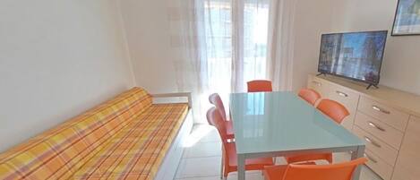 Una habitación con sofá, mesa de comedor con sillas, televisor sobre un mueble bajo y ventana con cortinas.