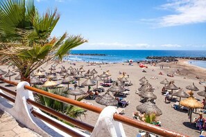 Een strand met rieten parasols en palmbomen, een heldere lucht en een rustige zee.