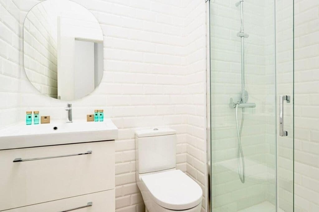 A bathroom with a white tiled wall, a round mirror, a white sink with toiletries, a toilet, and a glass-enclosed shower.