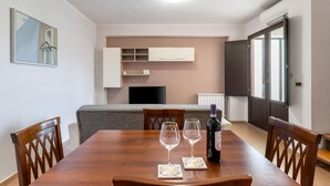 Ein Essbereich mit einem Holztisch, zwei Weingläsern und einer Weinflasche. Im Hintergrund befinden sich eine graue Couch und eine weiße Kommode.