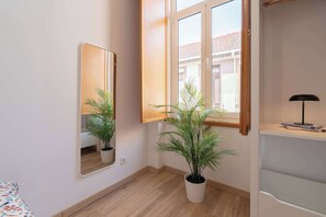 Ein Zimmer mit Holzboden, einem großen Spiegel, einer Topfpflanze, einem Fenster mit Blick auf Gebäude und einem Regal mit Lampe und Buch.