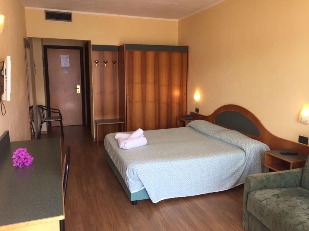 A hotel room with a bed, a desk, a chair, a wardrobe, and a small table.