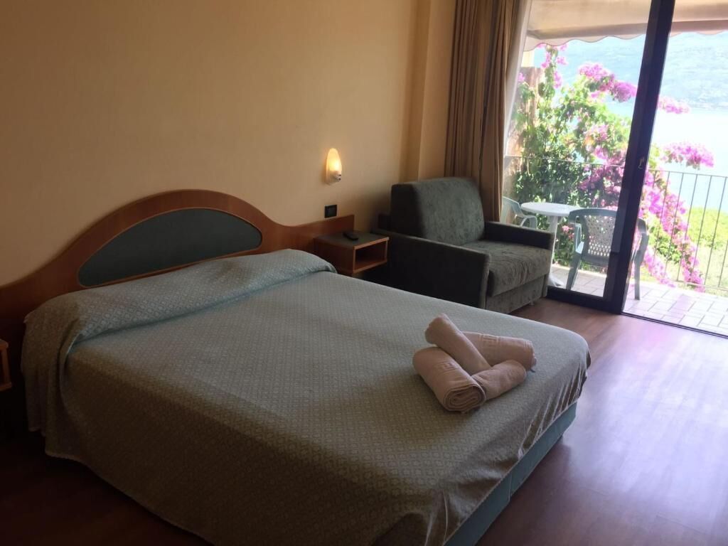 A hotel room with a bed, a chair, a small table, and a balcony with flowers.
