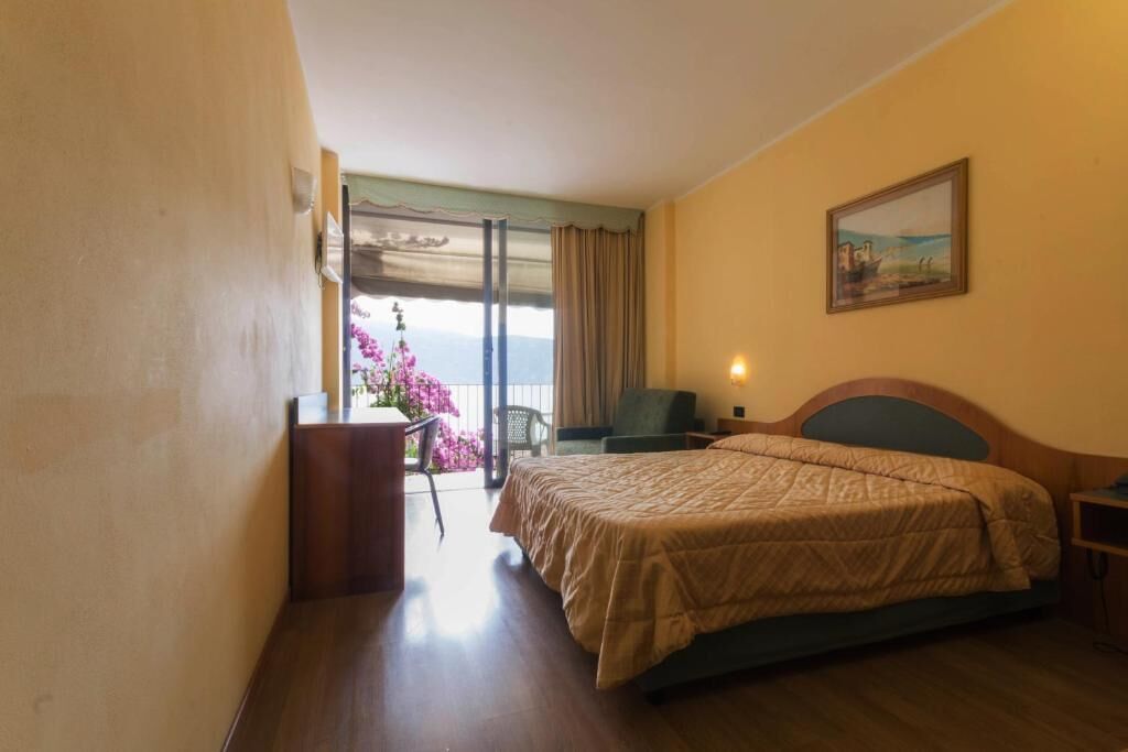 A hotel room with a bed, a desk, a chair, a painting, and a view of the sea.