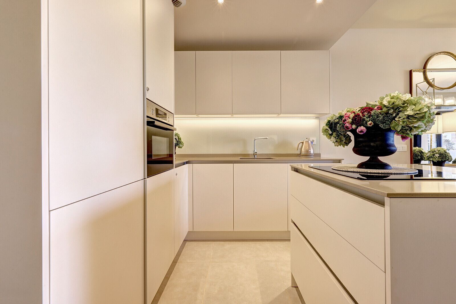 A modern kitchen with white cabinetry, a built-in oven, a stainless steel sink, and a vase with flowers on the counter.