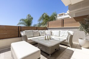 Eine Terrasse mit einer Rattan-Couch, einem weißen Hocker und einem Glastisch mit Kerzen.