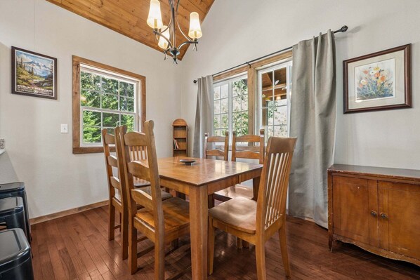 A dining area with a wooden table and chairs, large windows, a chandelier, and a cabinet. - Forest Ridge Chalet | Tesla EV, Hot Tub (Packwood)