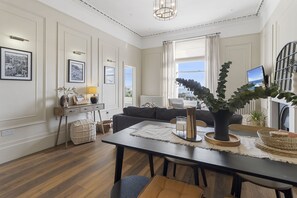 A modern living room with a sofa, dining table, and a large window with curtains. - Sea Breeze is a stylish two-bedroom luxury coastal apartment (Teignmouth)