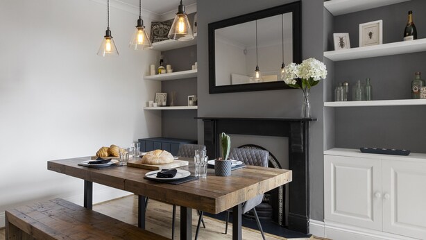 Un espace repas comprenant une table en bois, des chaises, des lampes suspendues, une cheminée et des étagères où sont disposés divers objets.
