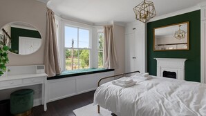 Une chambre à coucher avec un grand lit, un coin fenêtre aménagé en siège, une commode blanche, un pouf vert et une cheminée.