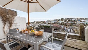 Un espacio de comedor al aire libre con una mesa, sillas y una sombrilla. Hay copas de vino, una botella y una cesta de pan sobre la mesa. Se ve una vista de un pueblo con casas coloridas al fondo.