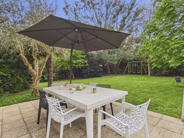 A patio with a table and chairs, an umbrella, and a swing set in the background. - Comfortable house for 9, with a garden and trees (Pléneuf-Val-André)