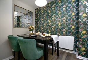 Zona de menjador amb taula preparada per a quatre, cadires tapissades de verd, un ram de flors i un paper pintat amb disseny.