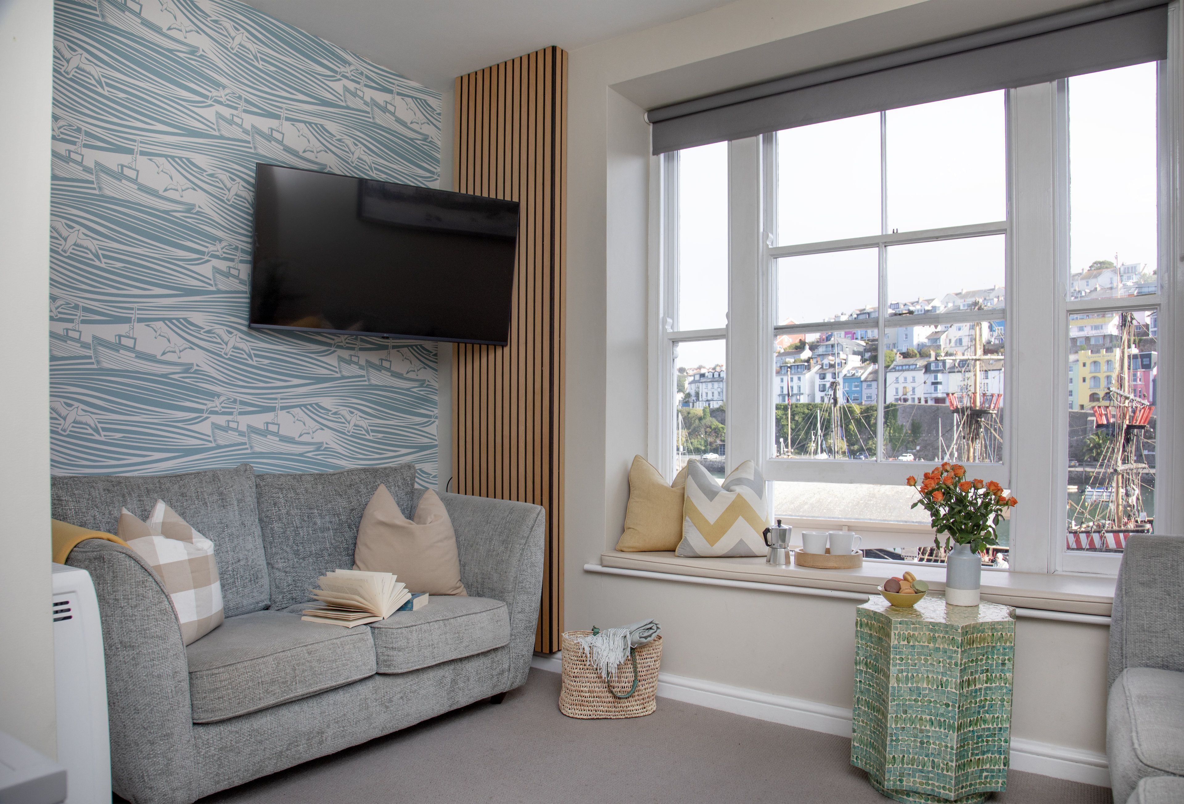 A living room with a grey sofa, a flat-screen TV mounted on the wall, a window with a view of a cityscape, and a small side table with a vase of flowers.