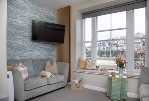 A living room with a grey sofa, a flat-screen TV mounted on the wall, a window with a view of a cityscape, and a small side table with a vase of flowers. - Seven - La Petite Maison is a stylish, dog-friendly holiday home in Brixham (Brixham)