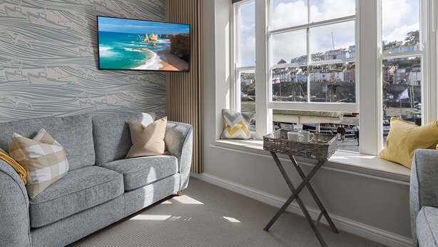 Ein Wohnzimmer mit grauem Sofa, einem an der Wand montierten Fernseher, einem Fenster mit Blick auf eine Küstenstadt und einem kleinen Tisch mit einem Korb.