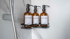 Casa de banho com parede de mármore, chuveiro e três frascos de produtos de higiene pessoal numa prateleira.