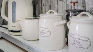 Uma bancada de cozinha com painel de fundo de pedra, uma cafeteira e dois recipientes de cerâmica branca com as etiquetas 'Café' e 'Chá'.