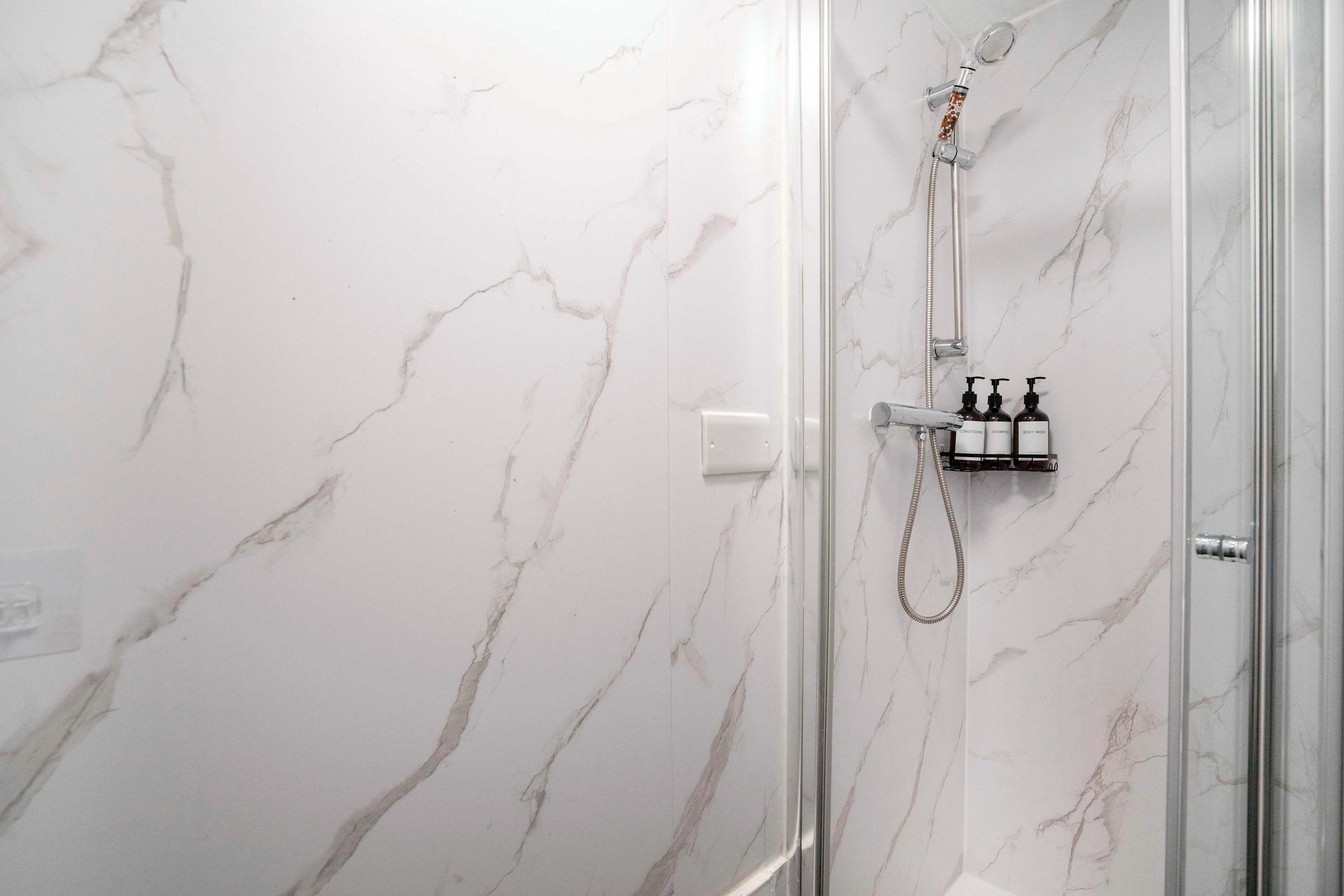 Een douche met een glazen deur, marmeren wanden en een handdouche.