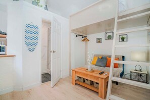 A modern living room with a wooden coffee table, a sofa with cushions, a white bookshelf, and a decorative zigzag wall art piece. - West Kensington Mezzanine Studio | 3-Min Tube | Sleeps 3 (Greater London)